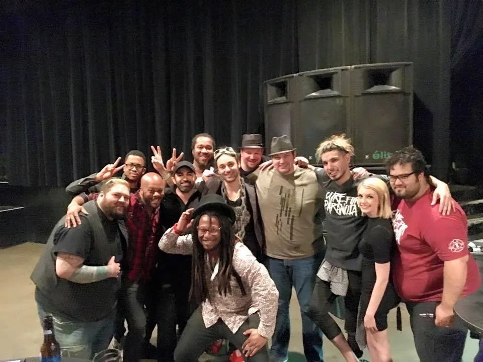 A dozen musicians and crew crowded together in front of the stage at the end of the night, PA stacks and a dark curtain behind.