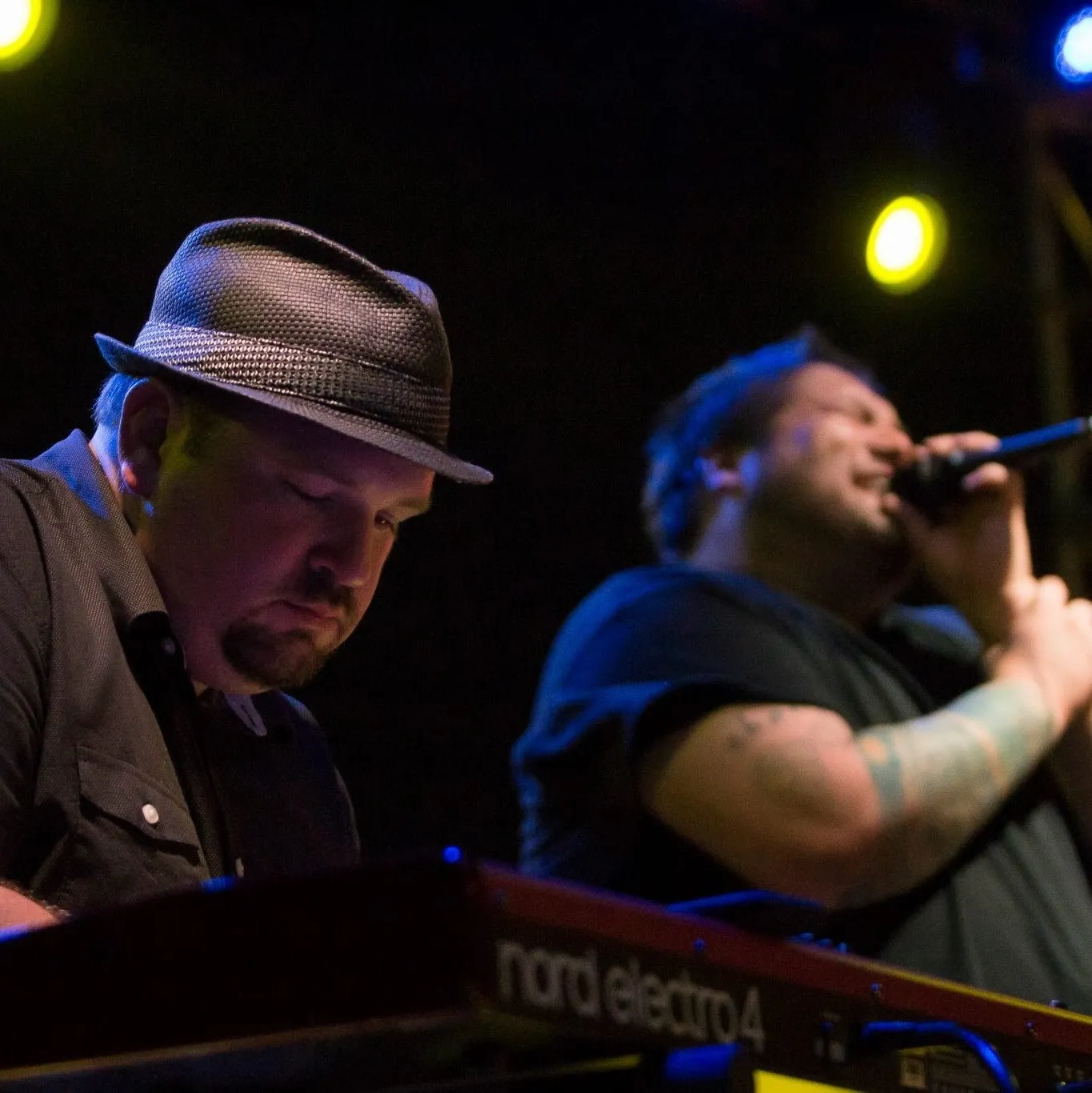 At a Nord Electro in a fedora, head down over the keys, a singer at the microphone alongside under blue and yellow light.