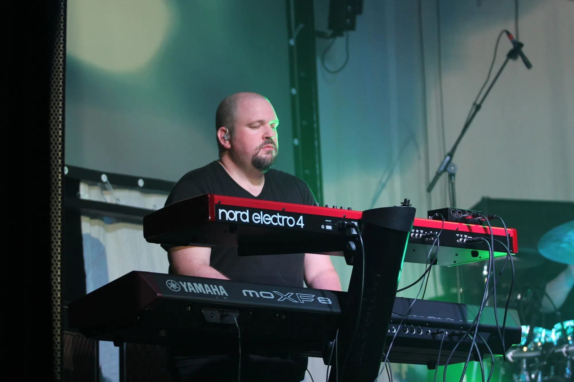 At a two-tier keyboard rig on stage, lit green, a Nord Electro over a Yamaha stage piano.