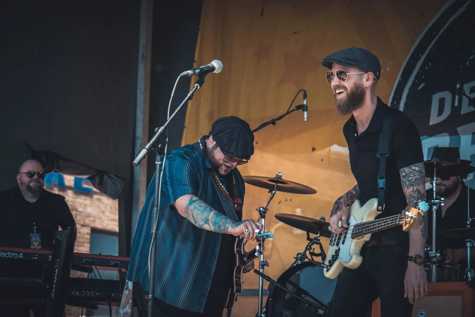 On an outdoor festival stage: at the keys on the left, laughing, while Paco Estrada tunes and Brandon Callies grins over his bass.