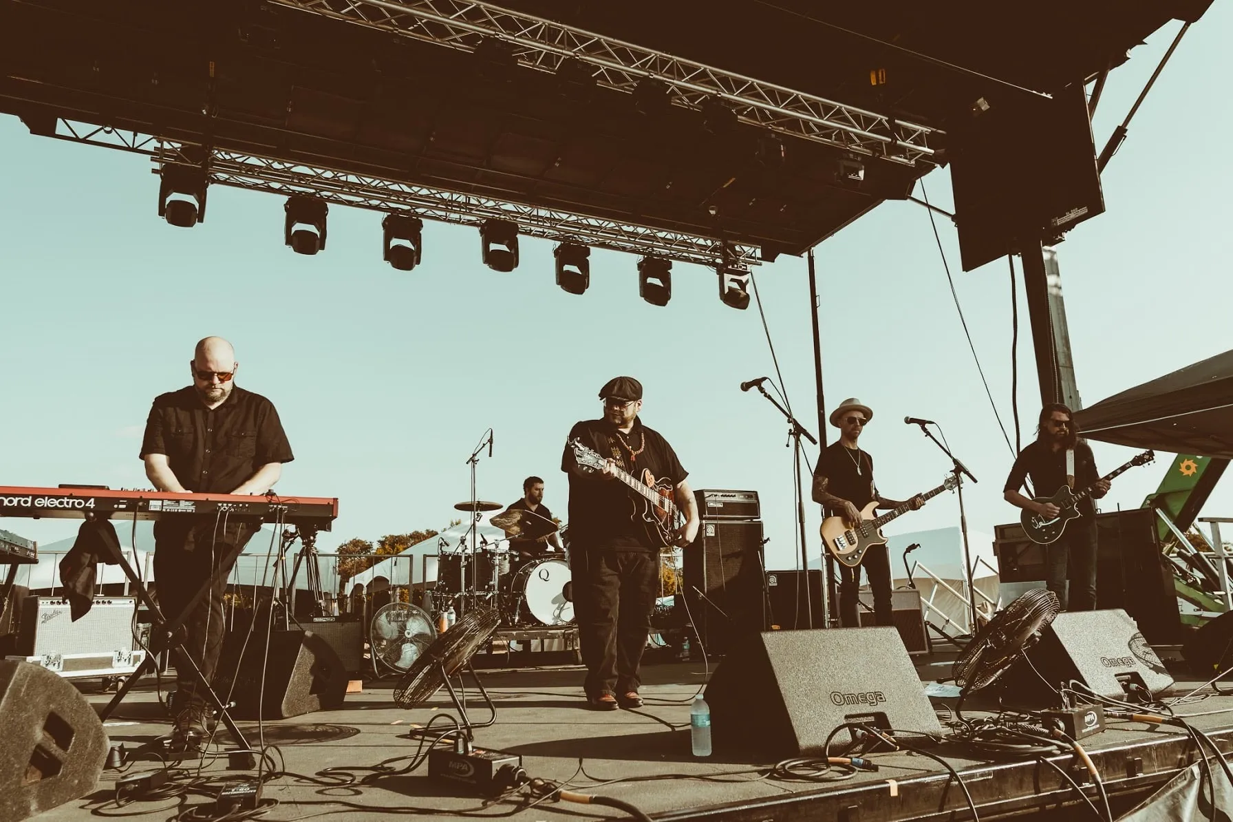 A daylight festival stage under a lighting truss, a Nord Electro at the front left, the band spread across the stage.