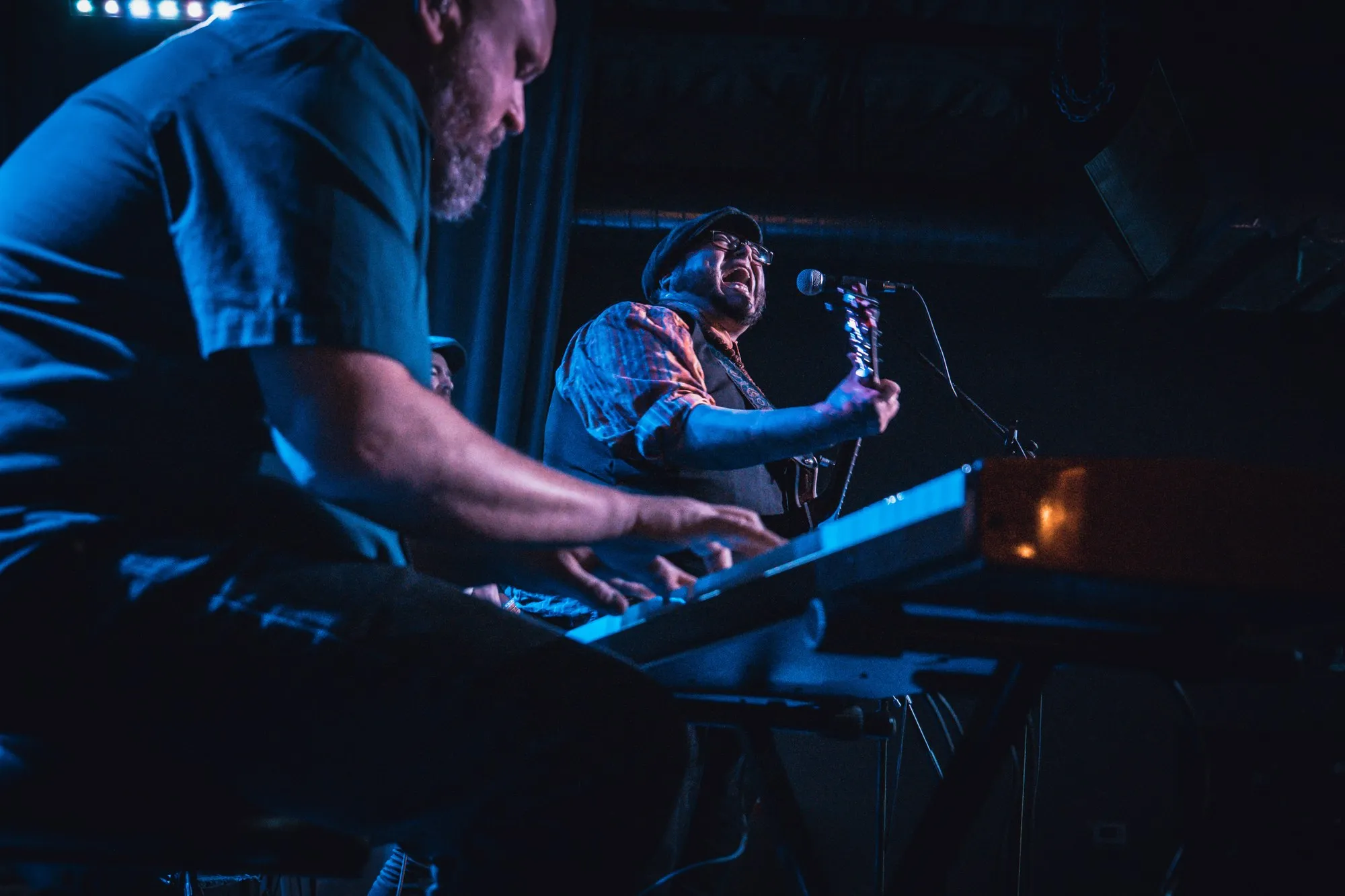 Hands on the keys in blue stage light, a singer with a guitar behind.