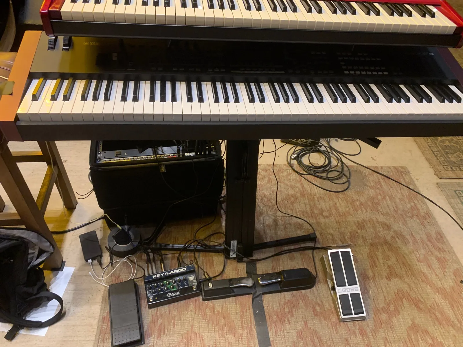 Looking down on a two-tier keyboard rig: a red Nord above a Yamaha stage piano, with a Radial Key-Largo mixer, sustain, expression and volume pedals on the floor.