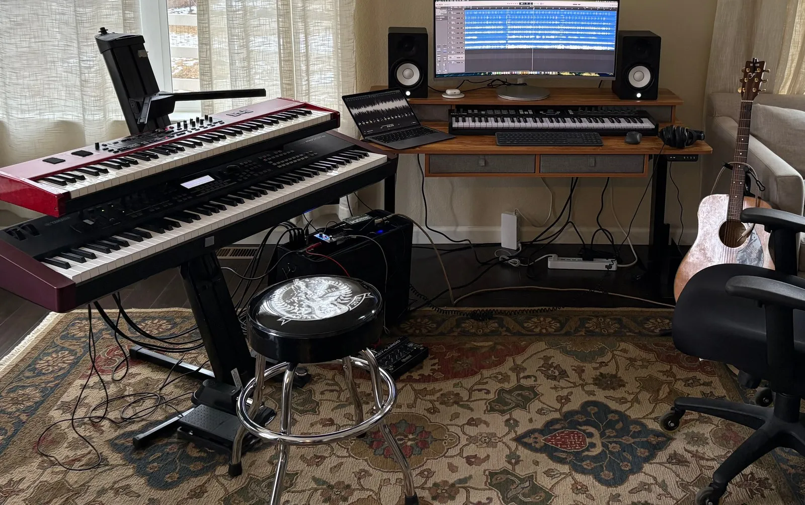 A home studio: the same two keyboards on a stand beside a desk with a laptop, a wide monitor showing a recording session, studio monitors, a controller keyboard and an acoustic guitar.