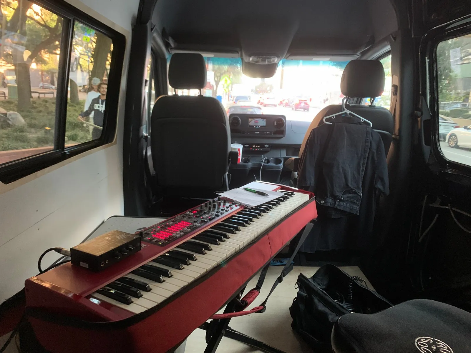 A red Nord keyboard on a stand inside a van, a handwritten chart and pen resting on it, a queue of people visible through the window.